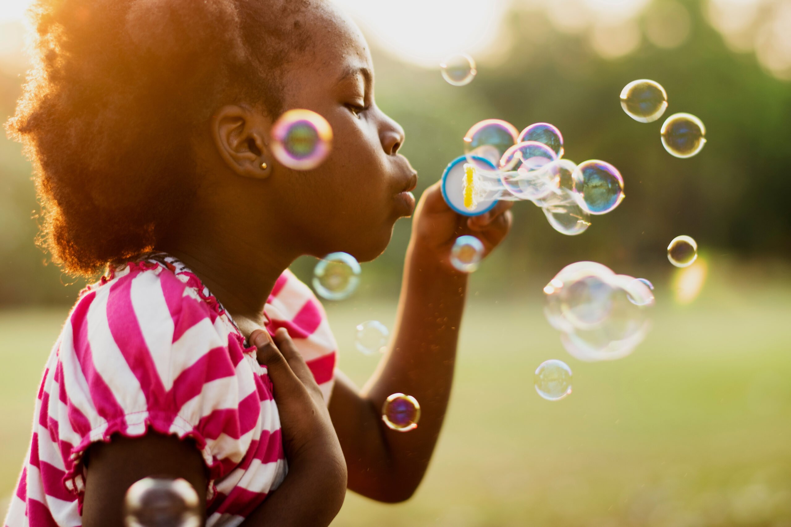 Cute Black girl in pink striped shirt blowing bubbles in the grass