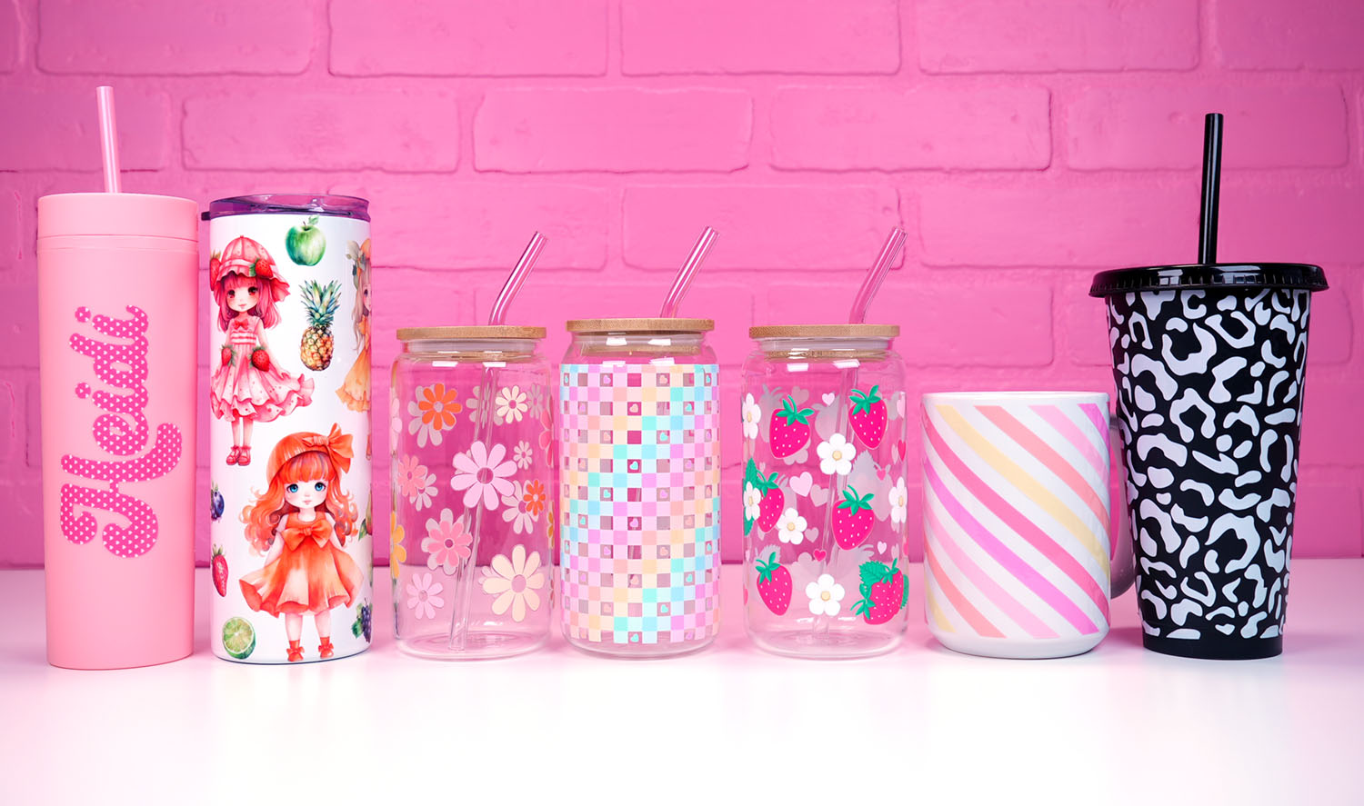 Assorted colors and sizes of tumblers and mugs with UV DTF wraps on a pink brick background